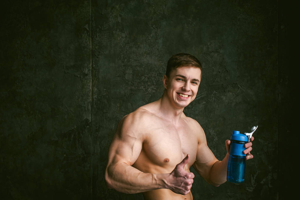 Mladá sexy mužů kulturista sportovec, studio podkroví portrét na pozadí stylizovaného zdi, guy model holé torzo nápoje vodou z láhve sportovní modré barvy po tréninku - Fotografie, Obrázek