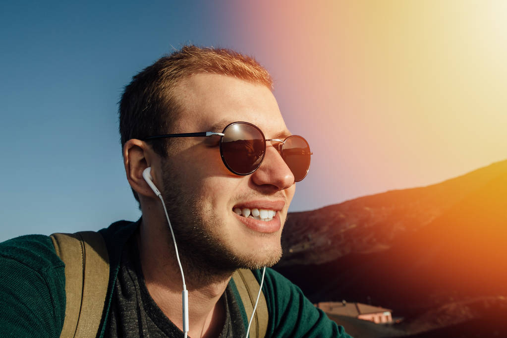 Портрет молодого чоловіка в сонцезахисних окулярах слухати музику в навушниках і посміхатися на заході сонця
 - Фото, зображення