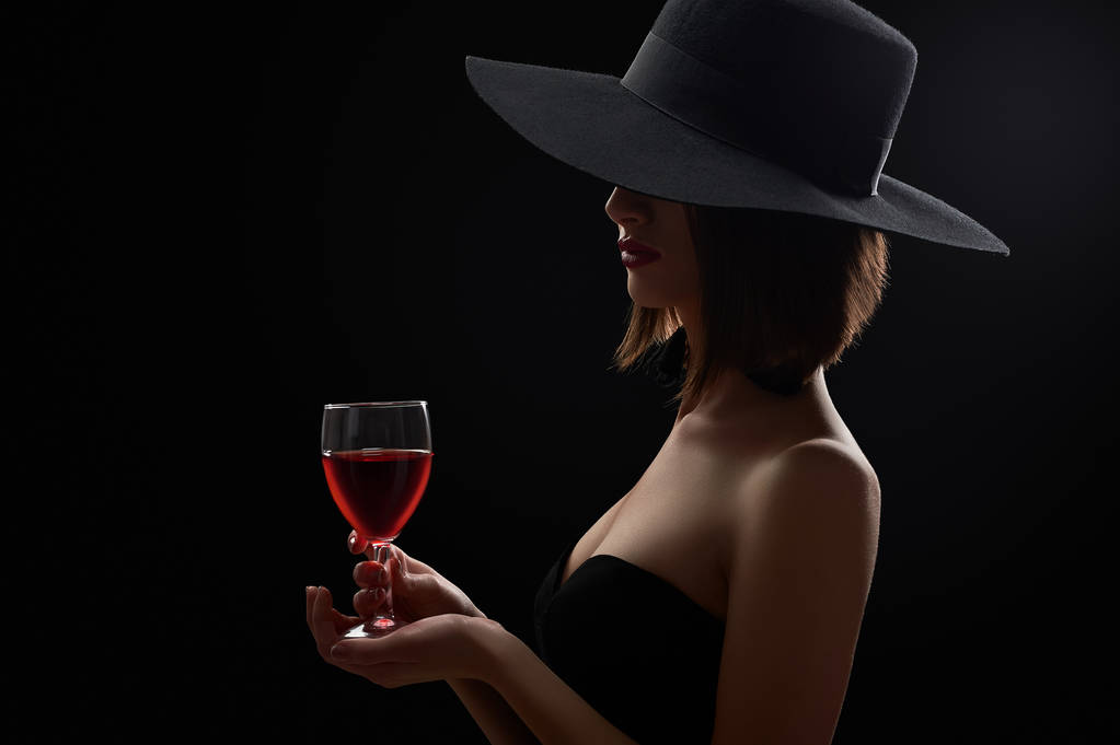 Elegante mujer misteriosa en un sombrero sosteniendo una copa de vino tinto en
 - Foto, Imagen