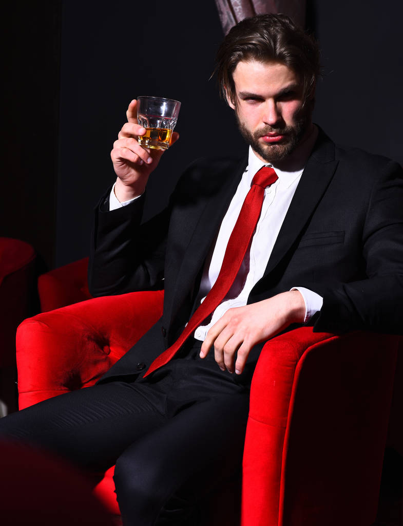 γενειοφόρος άνδρας, επιχειρηματίας κρατώντας ένα ποτήρι ουίσκι σε κόκκινη καρέκλα - Φωτογραφία, εικόνα