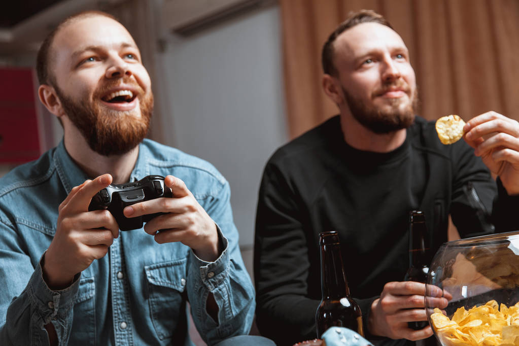 мужчины с бородой сидят дома на диване с пивом и джойстиками в руках, играя в компьютерные видеоигры
 - Фото, изображение