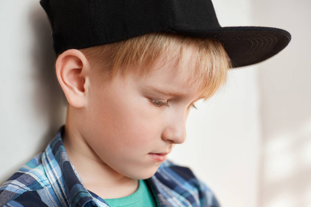 Portrait of adorable little boy with blond hair wearing stylish clothes and cap having thoughtful expression looking down standing near white wall. Beautiful and stylish child. Fashion and children. - Photo, Image