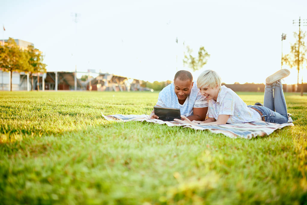 Casal de raça mista de millennial em um campo de grama olhando para um tablet digital e leitura para o seu próximo jornal da escola
 - Foto, Imagem