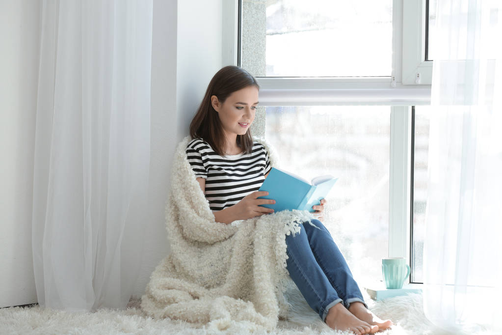 Νεαρή γυναίκα κάθεται στο παράθυρο και διαβάζοντας το βιβλίο στο σπίτι - Φωτογραφία, εικόνα