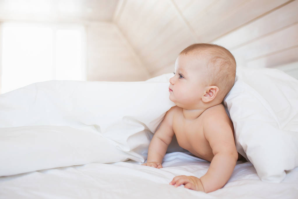 Pequeno bebê olha para um lado de um cobertor em uma cama branca
 - Foto, Imagem