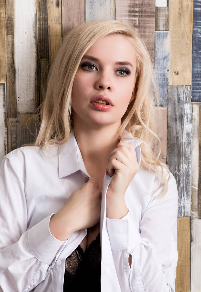 portrét atraktivní blond dívka stojící na dřevěnou zeď na pozadí. Má modré oči a oblečený v mans košile - Fotografie, Obrázek