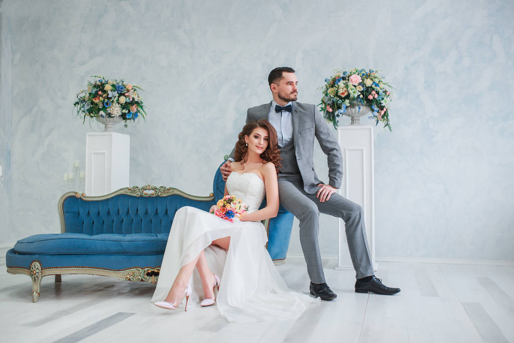 Bride in beautiful dress and groom in gray suit sitting on sofa indoors in white studio interior like at home. Trendy wedding style - Photo, Image
