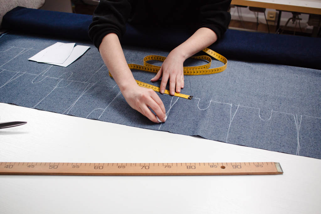 Руки швачки для вирізання джинсової тканини з візерунком ножиць та вимірювальною стрічкою на білому столі
 - Фото, зображення