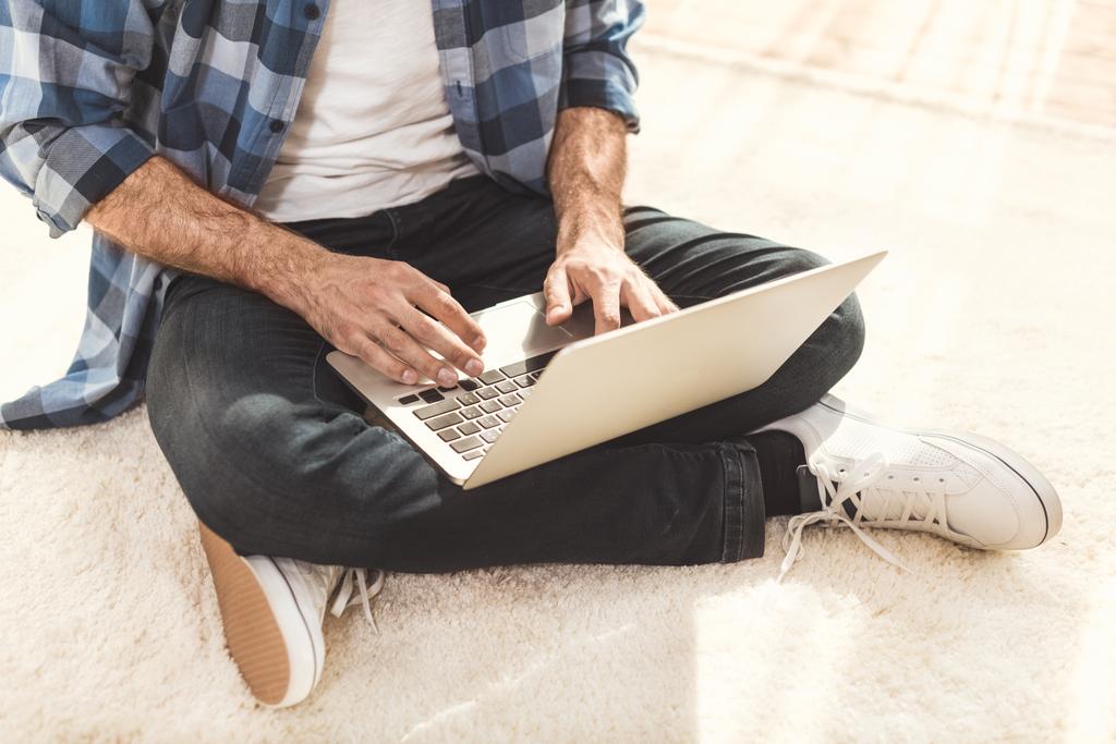 Чоловік сидить на килимі і друкує на ноутбуці
 - Фото, зображення