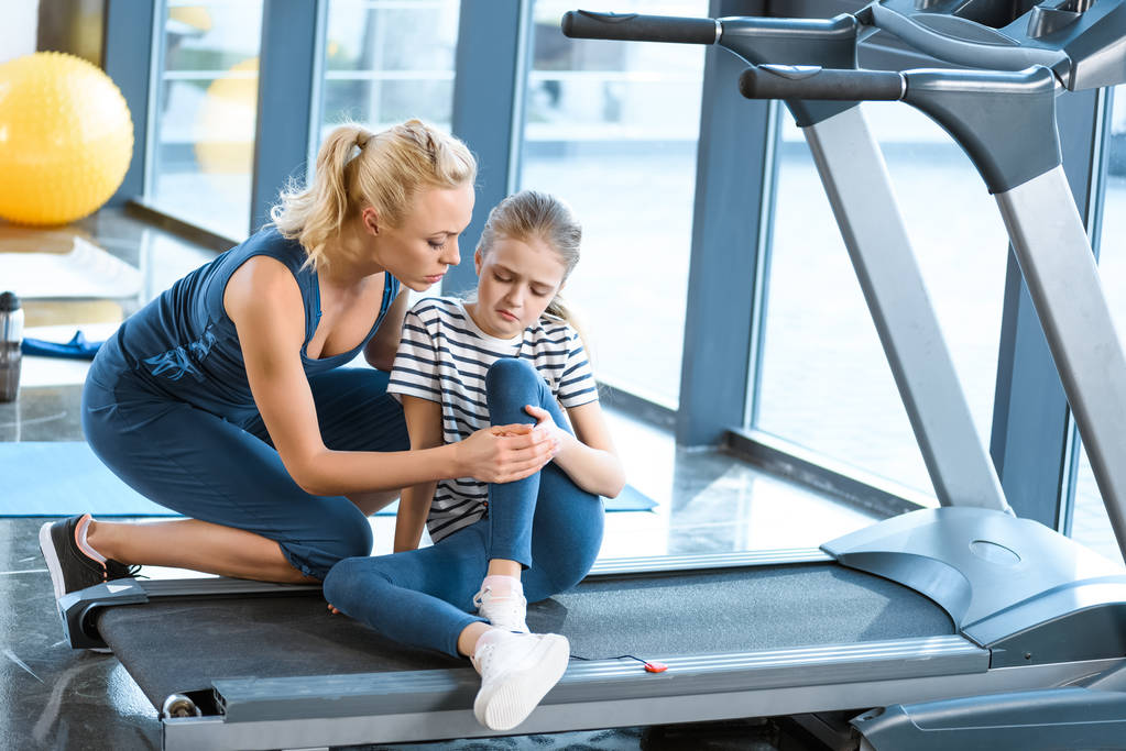 Woman helping girl injured knee sitting on treadmill - Photo, Image