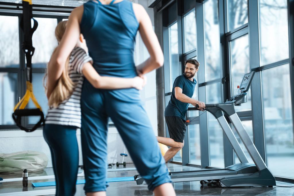 Frau mit Mädchen sieht gutaussehenden Mann beim Training auf dem Laufband im Fitnessstudio an - Foto, Bild