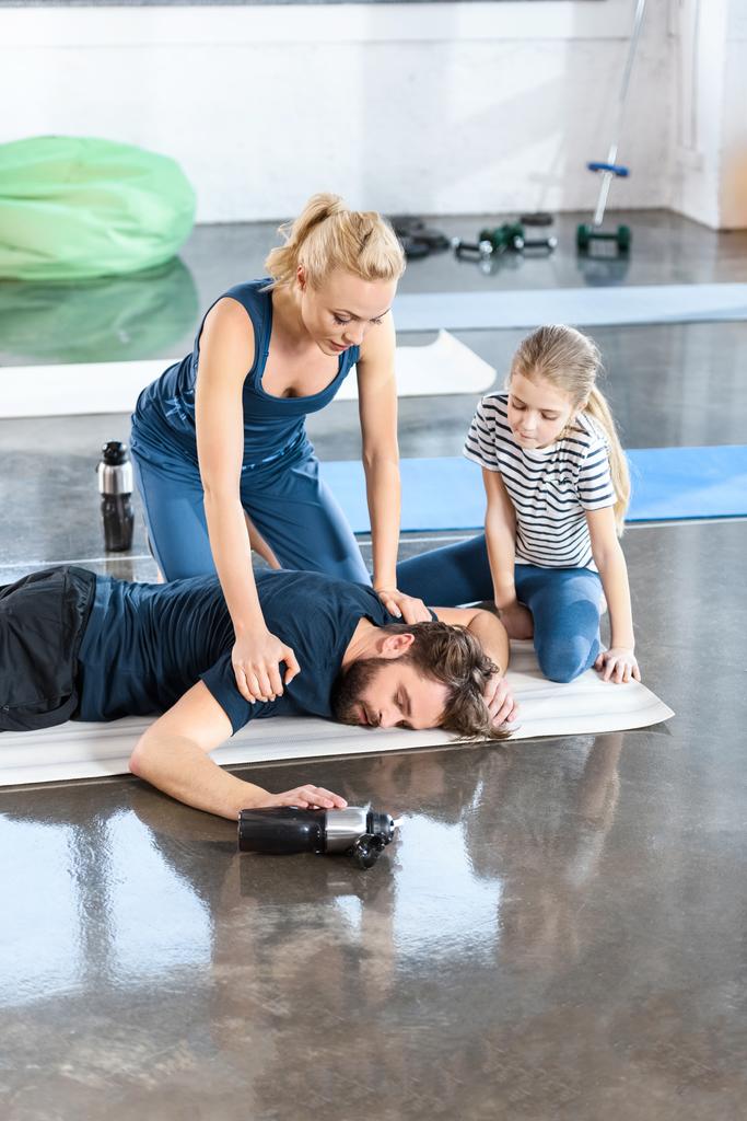 Жінка з дівчиною допомагає втомленому чоловікові лежати на килимку в спортзалі
 - Фото, зображення