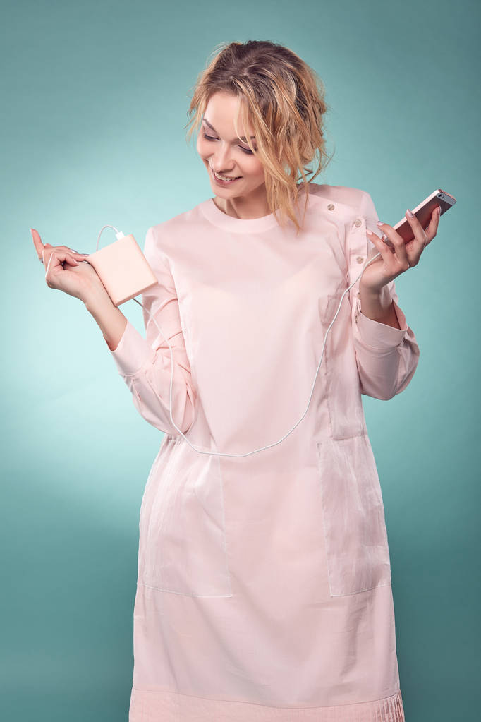 Giovane donna bionda romantica con i capelli ondulati in abito rosa chiaro che tiene smartphone rosa in studio su sfondo turchese
 - Foto, immagini