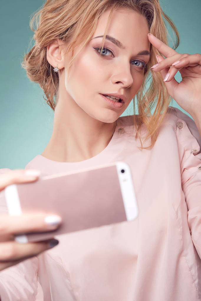 Крупный план портрет молодой романтичной улыбающейся блондинки с волнистыми волосами в светло-розовом платье, смотрящей на розовый смартфон в студии на бирюзовом фоне
 - Фото, изображение