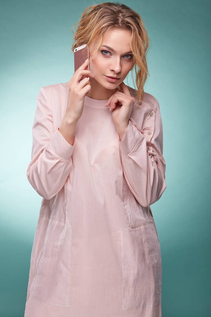 Young beautiful blonde woman with wavy hair in light pink dress talking by pink smartphone and looking serious in studio on turquoise background - Photo, Image