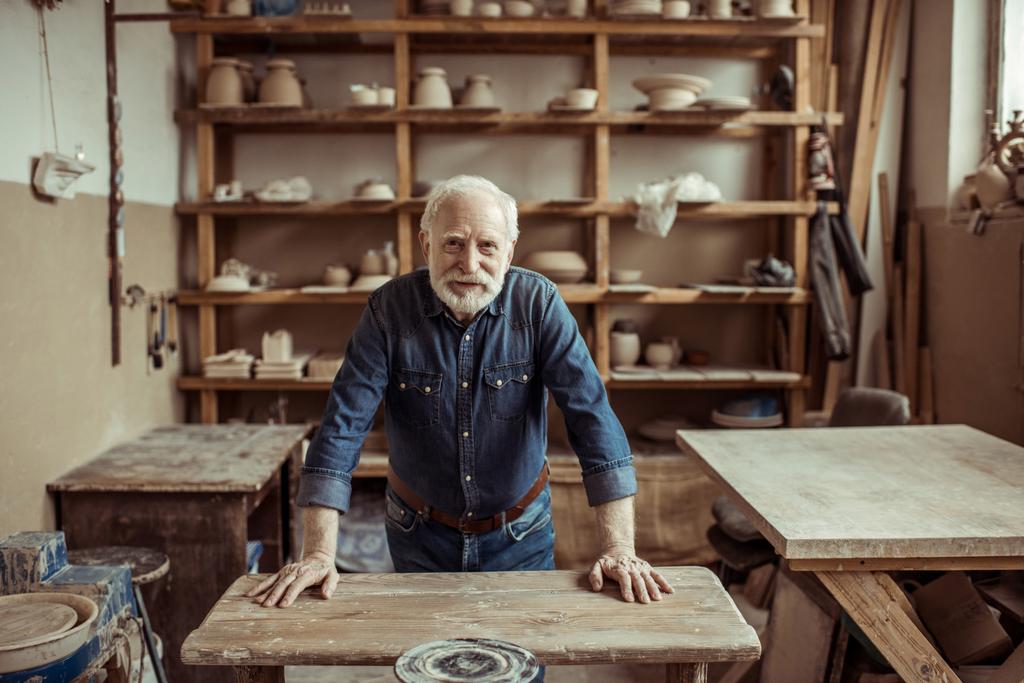 Vista frontale del vasaio anziano in piedi e appoggiato sul tavolo contro scaffali con ceramiche in officina
 - Foto, immagini