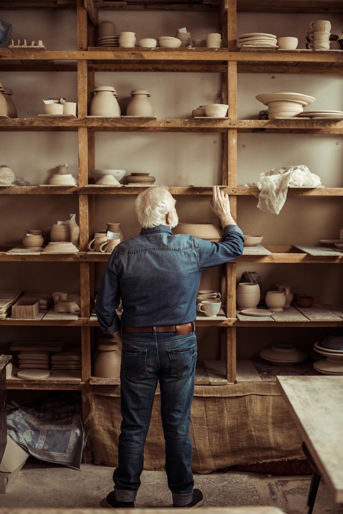 Rückansicht eines älteren Töpfers, der neben Regalen mit Töpferwaren steht und in der Werkstatt nach etwas sucht - Foto, Bild