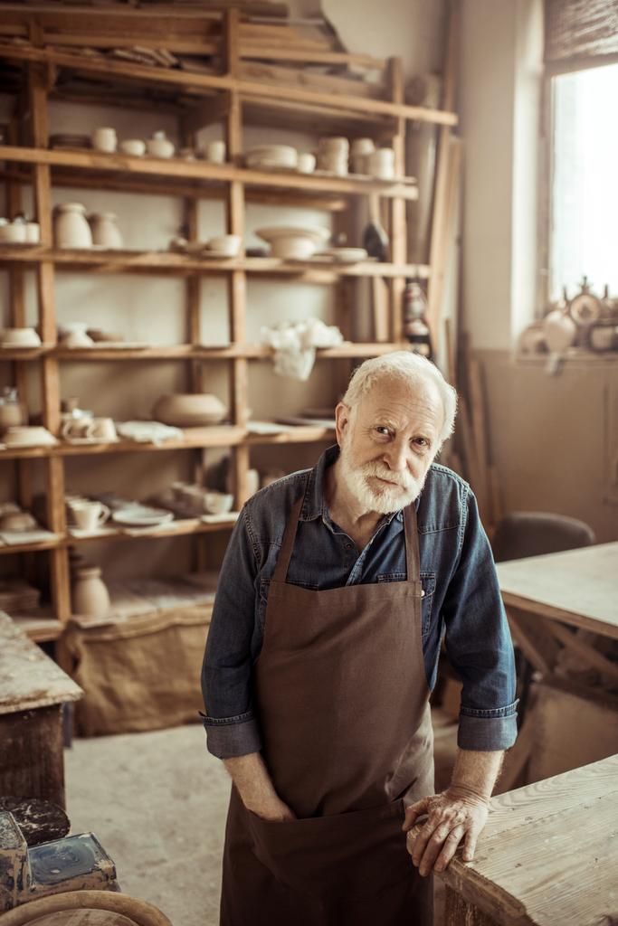 Frontansicht des Senior-Töpfers in Schürze stehend und auf einem Tisch gegen Regale mit Töpferwaren in der Werkstatt gelehnt - Foto, Bild