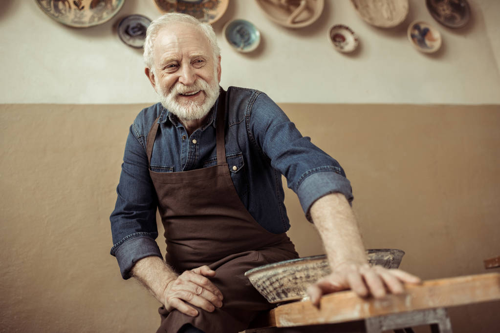Potier senior dans tablier assis sur la table contre le mur avec des articles de poterie suspendus
 - Photo, image