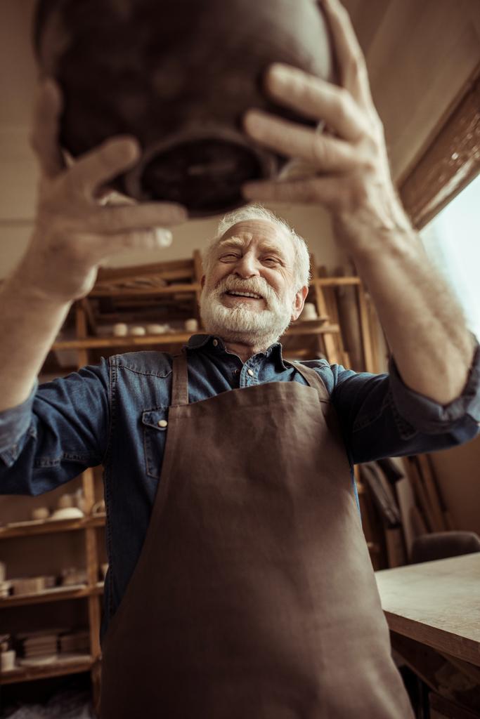 Старший гончар в фартуке рассматривает керамическую миску в мастерской
 - Фото, изображение