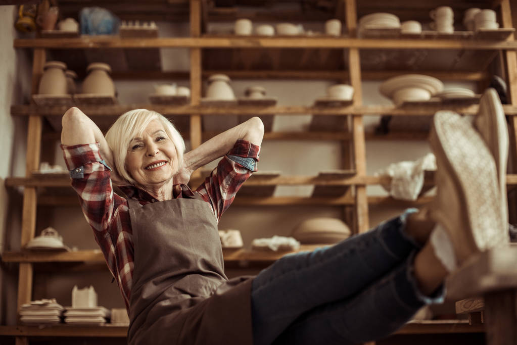Vista frontal da mulher idosa sentada na cadeira com as pernas na mesa contra prateleiras com produtos de cerâmica
 - Foto, Imagem