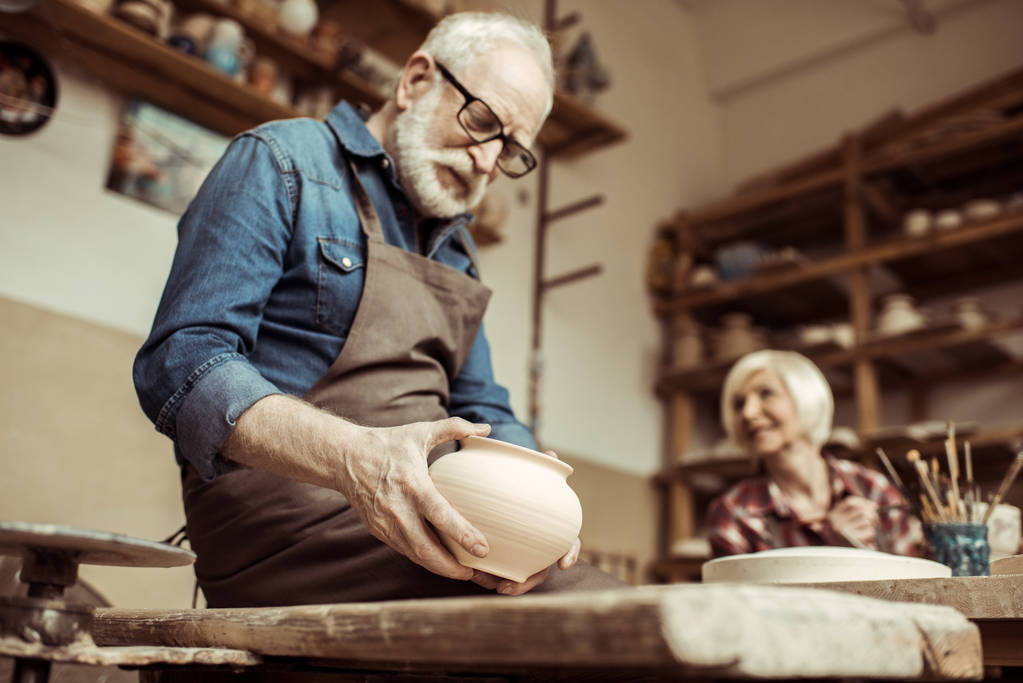 Starszy potter w fartuch i okulary badanie miski ceramiczne z kobieta na tle - Zdjęcie, obraz