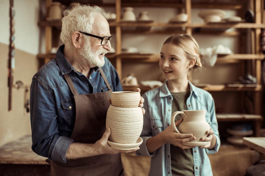 Внучка и дедушка держат и осматривают глиняные товары
 - Фото, изображение