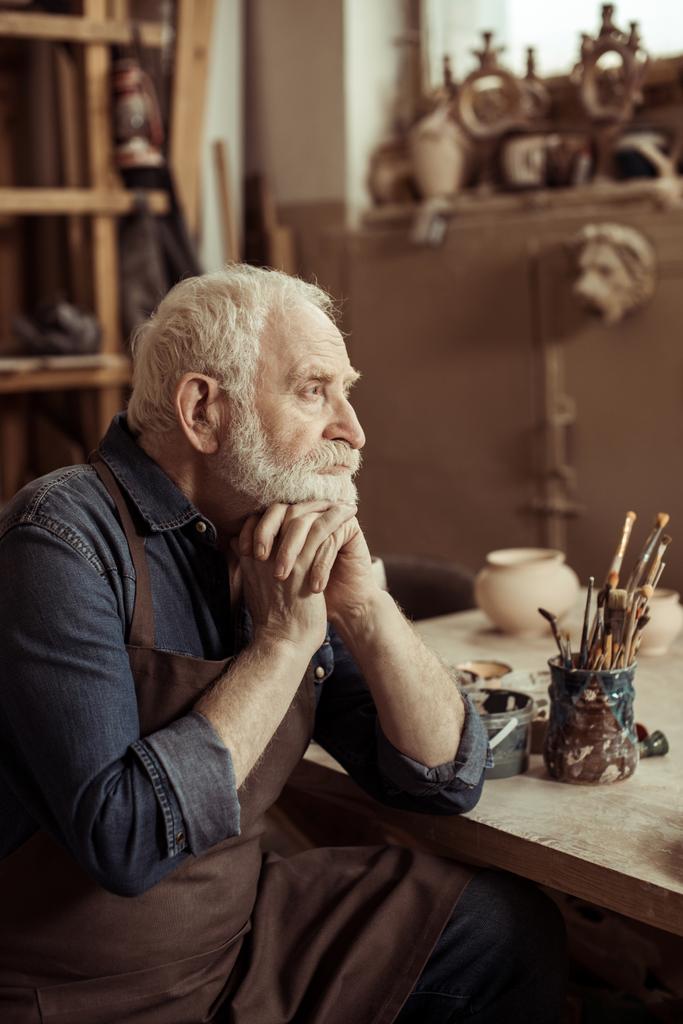 Senior potter in apron sitting at table and daydreaming at manufacturing - Photo, Image