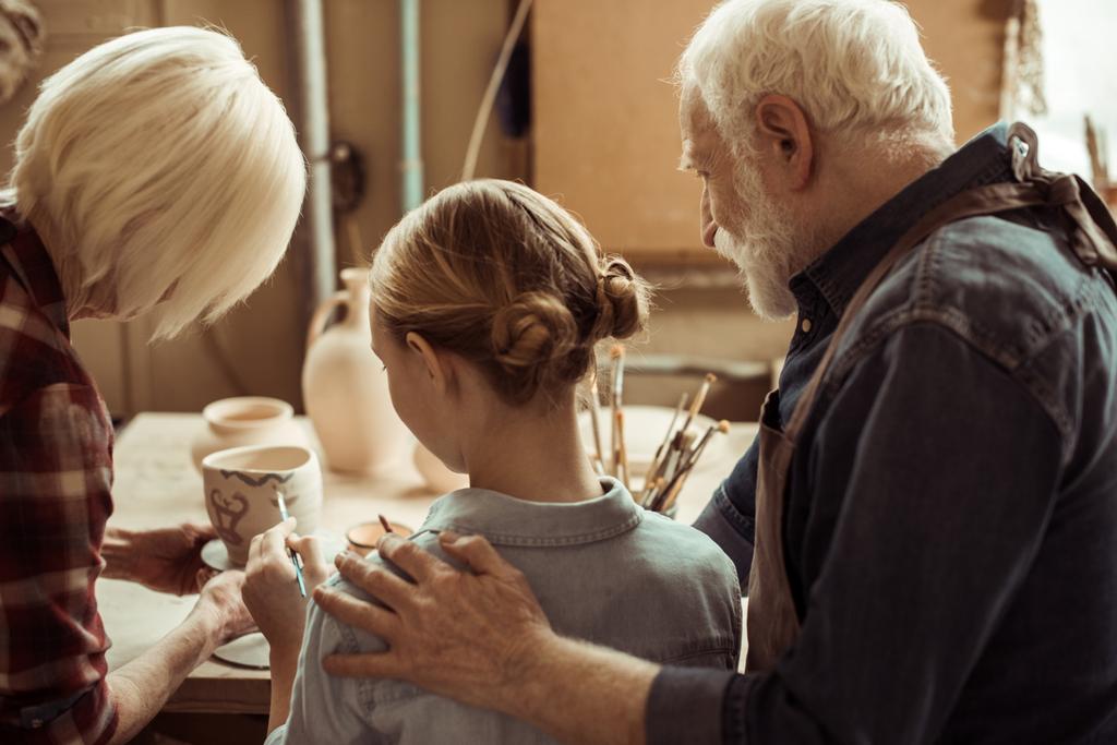 Οπίσθια όψη της κορίτσι ζωγραφική πήλινο δοχείο και τους παππούδες τους βοηθώντας στο εργαστήριο - Φωτογραφία, εικόνα