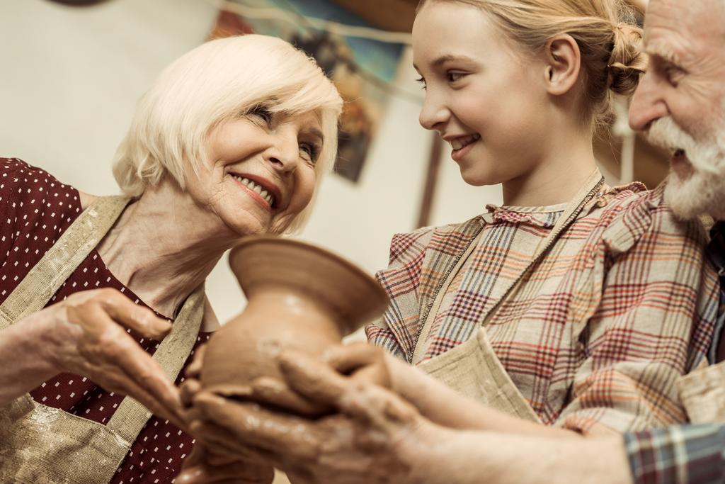 Бабушка и дедушка с внучкой держат глиняную вазу в мастерской
 - Фото, изображение