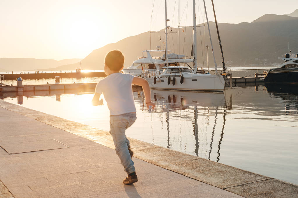 Poika juoksee lähellä jahteja satamassa auringonlaskun taustalla merellä
,  - Valokuva, kuva