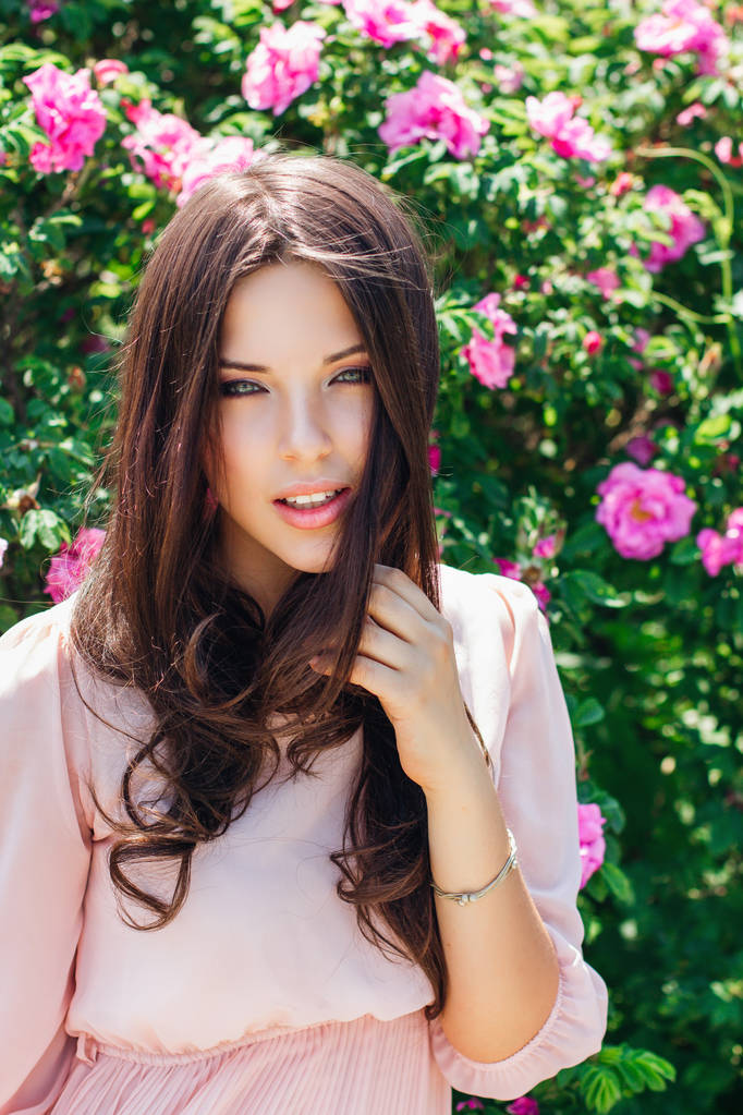 Hermosa joven con el pelo largo y rizado posando cerca de rosas en un jardín. El concepto de publicidad de perfumes
. - Foto, Imagen