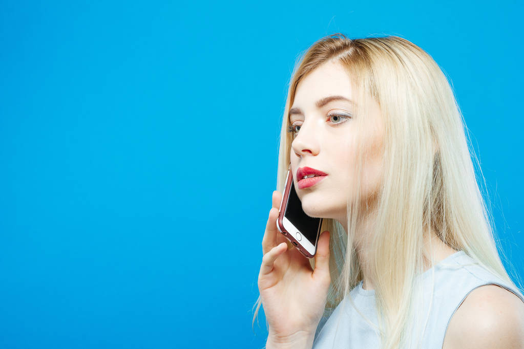 Seitenansicht der niedlichen Blondine im Kleid, die jemanden mit ihrem Smartphone auf blauem Hintergrund anruft. attraktives Mädchen mit Handy im Studio. - Foto, Bild
