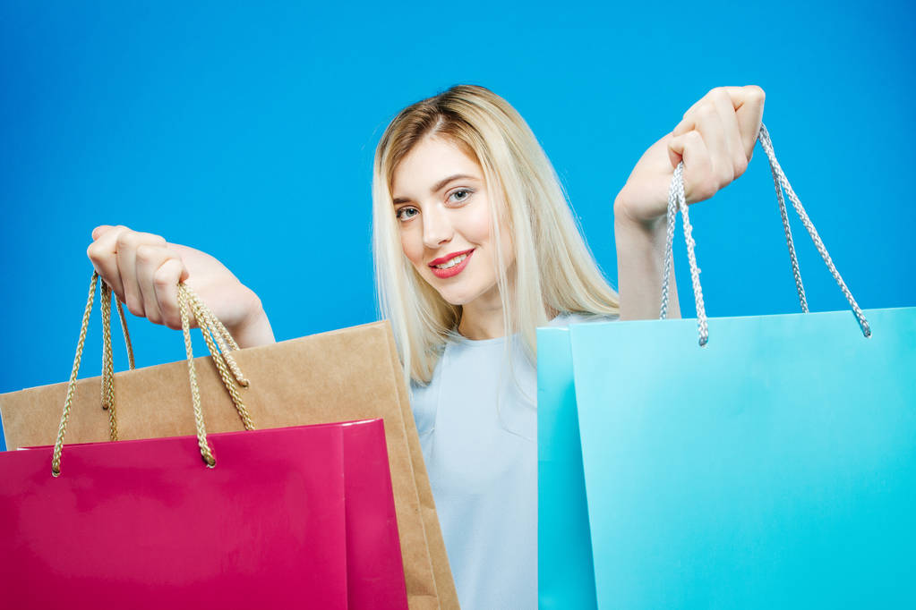 Nosit šaty roztomilé ženské Shopper je drží nákupní tašky na modrém pozadí. Šťastná dívka s vlasy Milano a okouzlující úsměv ve studiu. - Fotografie, Obrázek