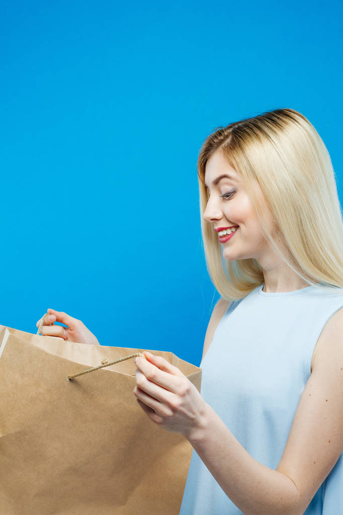 Retrato de la rubia sorprendida de aspecto agradable con bolsa de compras de papel en su mano posando sobre fondo azul. Chica de pelo rubio en el estudio
. - Foto, imagen