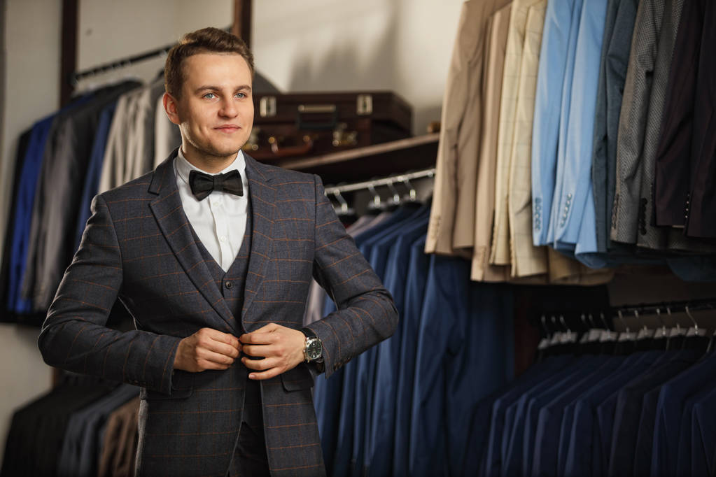 Businessman in classic vest against row of suits in shop. A young stylish man in a black cloth jacket. It is in the showroom, trying on clothes, posing. Advertising photo - Photo, Image