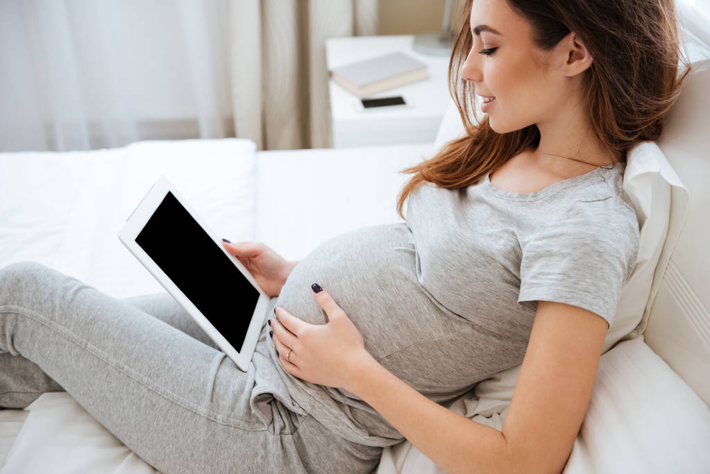 Sonriendo mujer joven embarazada mintiendo y utilizando la tableta de pantalla en blanco
 - Foto, Imagen