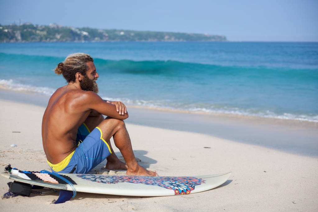 Homem encaracolado barbudo atraente com uma prancha de surf está sentado de costas em uma areia e olhando para o oceano
 - Foto, Imagem
