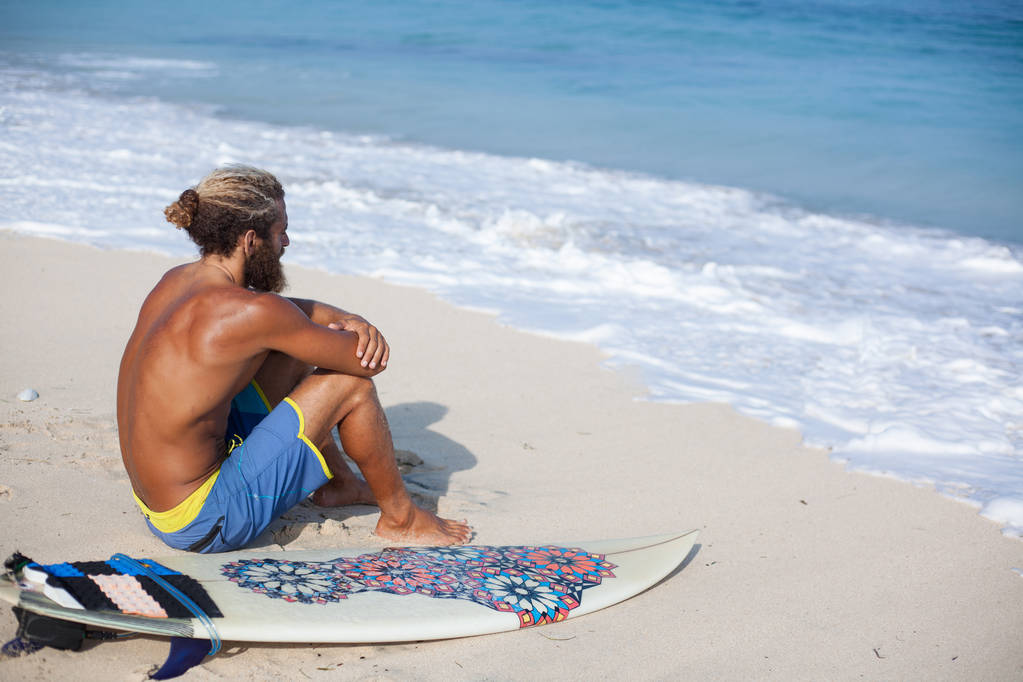 Çekici sakallı kıvırcık adam bir surfboard ile sırt üstü bir kum ile oturma ve okyanusa bakarak - Fotoğraf, Görsel