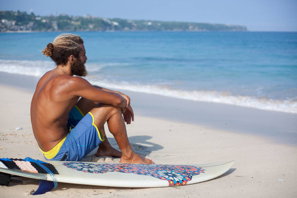muotokuva houkutteleva parrakas kihara mies surffilauta istuu selkä hiekalla ja katselee merelle
 - Valokuva, kuva