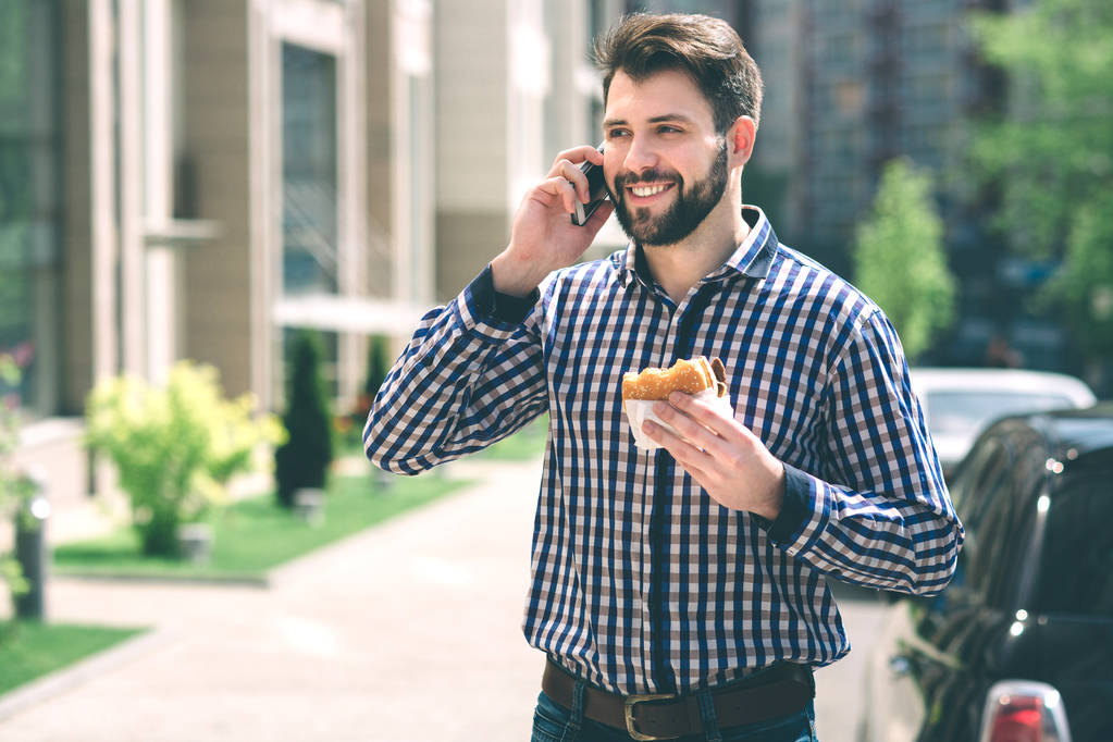 schöner sportlicher bärtiger dunkelhaariger Mann isst einen Hamburger und telefoniert - Foto, Bild