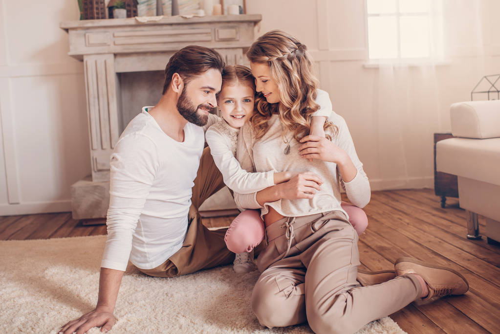 Счастливая молодая семья с одним ребенком, сидящим на ковре и обнимающимся дома
 - Фото, изображение