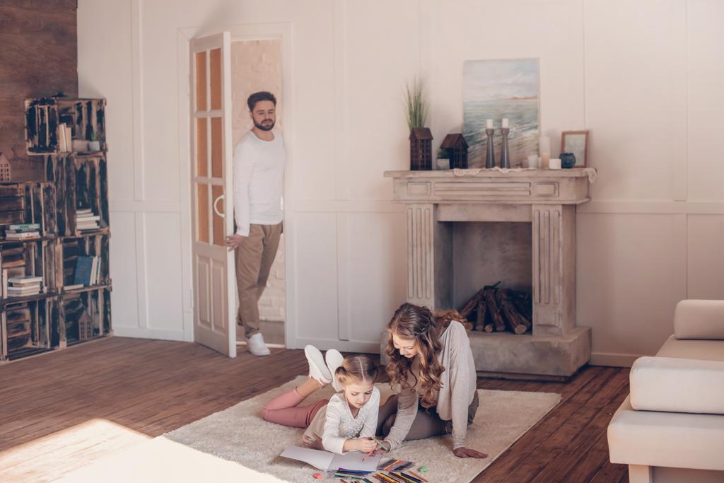 Homem em pé na porta e olhando para a mãe com a filha desenhando no chão
 - Foto, Imagem
