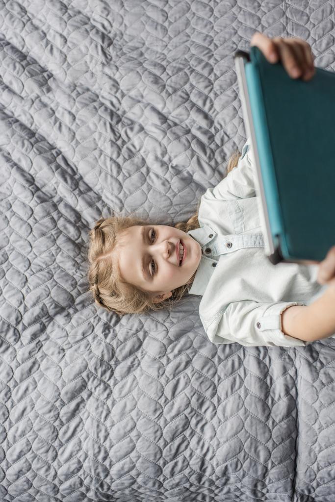 schattig meisje met behulp van digitale tablet en liggend op bed thuis - Foto, afbeelding