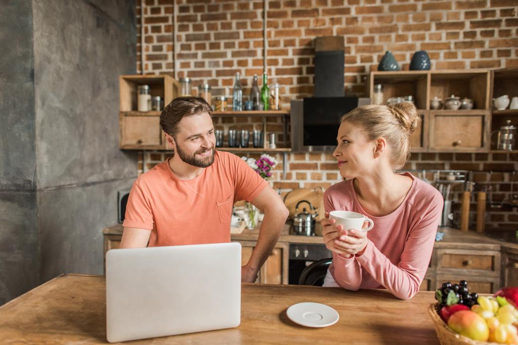 feliz jovem casal usando laptop na cozinha e sorrindo uns aos outros
 - Foto, Imagem
