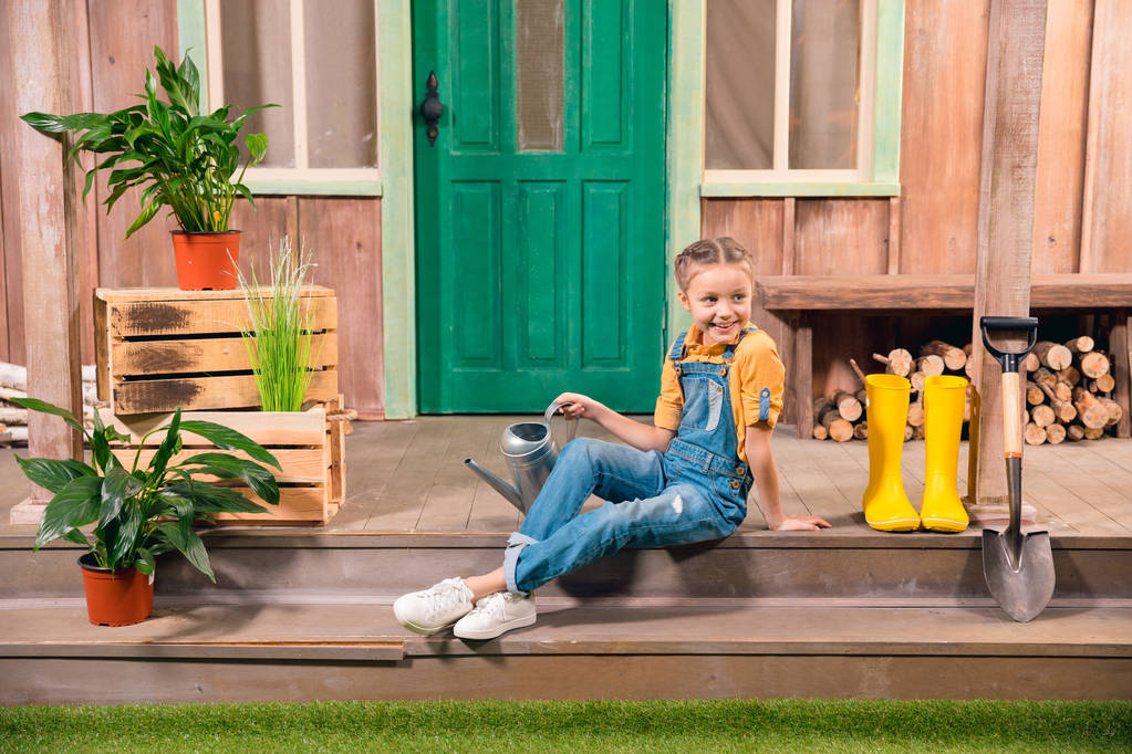 Suloinen hymyilevä pikkutyttö istuu kuistilla kastelukannulla ja katsoo pois
 - Valokuva, kuva