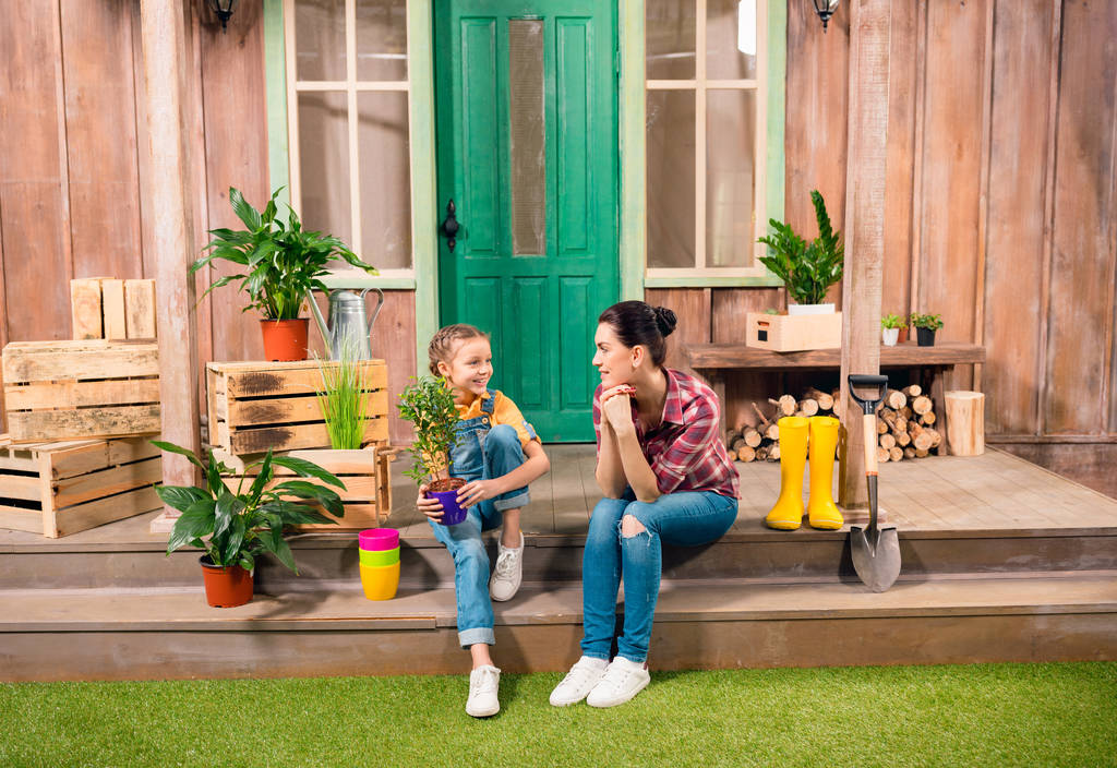 Feliz madre e hija con planta en maceta sentadas juntas en el porche y sonriendo entre sí
 - Foto, Imagen