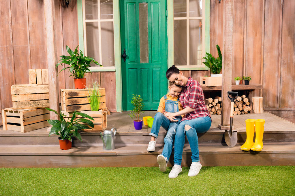 Feliz madre e hija sentadas y cogidas de la mano en el porche con plantas en maceta
 - Foto, imagen