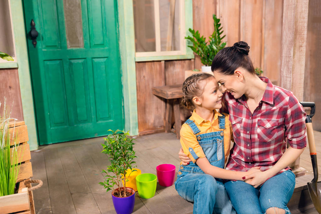 Feliz madre e hija sentadas y cogidas de la mano en el porche con plantas en maceta
 - Foto, imagen