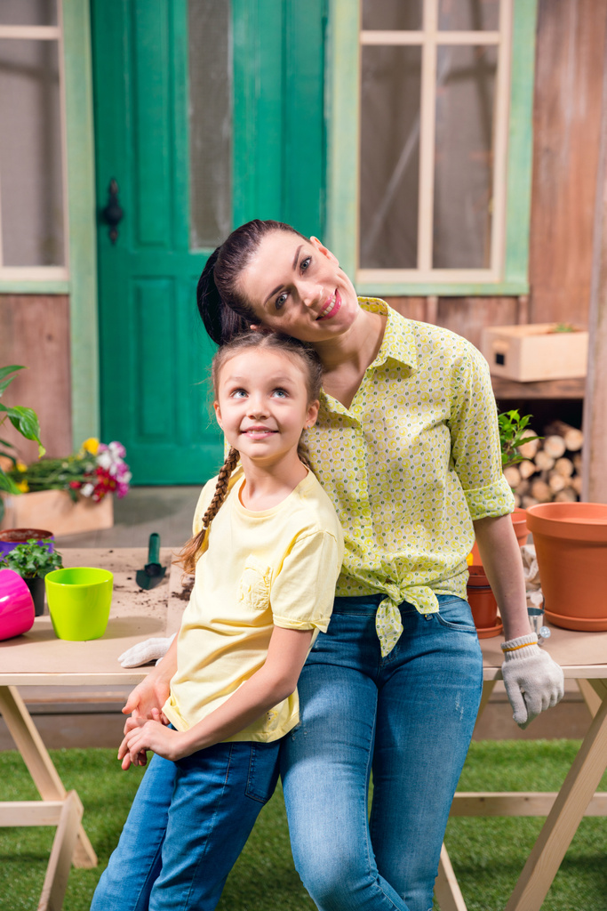 μητέρα και κόρη με φυτά και γλάστρες στέκεται και αγκάλιασμα στο τραπέζι στην βεράντα  - Φωτογραφία, εικόνα
